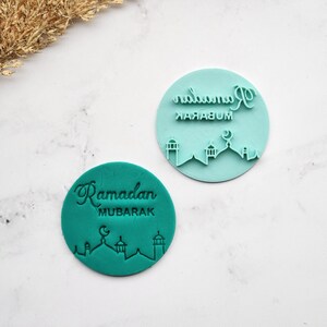 Peut inclure: Deux tampons à biscuits circulaires turquoise. L'un porte l'inscription "Ramadan MUBARAK" et une silhouette de mosquée, l'autre le même texte à l'envers. Ces tampons sont conçus pour faire des biscuits sur le thème du Ramadan.