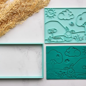 May include: Two teal-colored, square cookie cutters with an Easter theme. One cutter has a rectangular frame. The other two cutters feature a bunny, sun, clouds, trees, and a sign that says "Happy Easter".