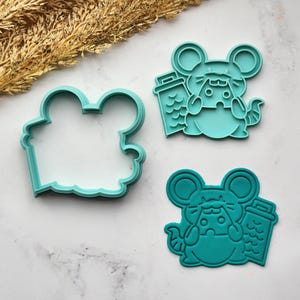 May include: Three teal-colored cookie cutters in the shape of a mouse. One cutter is the outline of the mouse shape, and the other two are the mouse design with a small container. The cutters are on a white marble surface with dried wheat.
