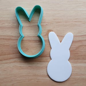 May include: A teal plastic bunny-shaped cookie cutter next to a white bunny-shaped cutout, both on a wooden surface. The cookie cutter has a rounded body and long ears, perfect for Easter treats.