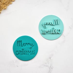 May include: Two teal-colored, circular stamps with the words "Merry Christmas" in a handwritten font. One stamp has the text in the correct orientation, and the other is a mirror image. A small Christmas tree graphic is included.