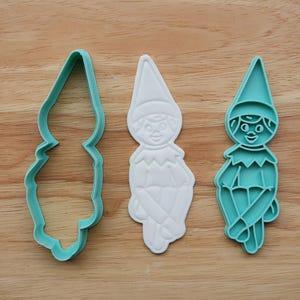 May include: Three elf-shaped cookie cutters. One is a teal outline, one is a white embossed elf, and the third is a teal elf with facial features and crossed legs. All are on a wooden surface.