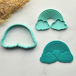 Può includere: Tre formine per biscotti a forma di arcobaleno color turchese. Due formine presentano un disegno ad arcobaleno con nuvole, e una è un semplice contorno. Le formine sono in plastica e si trovano su una superficie bianca.
