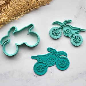 May include: Three teal-colored motorcycle-shaped cookie cutters. One is a solid outline, and the other two are detailed with wheels, seats, and handlebars. The cutters are arranged on a white marble surface, with dried wheat stalks in the background.