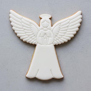 May include: A white angel-shaped cookie with detailed wing and halo designs. The cookie has a light brown edge and is set against a gray background. The angel is holding a small object in its hands.