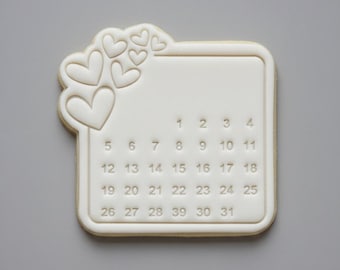 May include: A white cookie cutter in the shape of a calendar with a border of hearts. The calendar has numbers 1-31, perfect for baking themed treats. The cookie cutter is on a gray background.