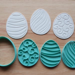 Könnte beinhalten: Eine Auswahl an Ausstechern in Eiform in Weiß und Türkis. Die weißen Ausstecher weisen verschiedene geprägte Designs auf, während die türkisfarbenen Ausstecher passende Designs haben. Die Ausstecher sind auf einer Holzoberfläche angeordnet.