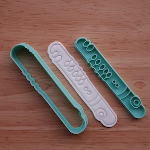 May include: Three plastic cookie cutters in the shape of a flute. One is a solid outline, one is white with details, and the other is teal with details. The cutters are on a wooden surface.