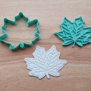 May include: Three maple leaf-shaped items on a wooden surface. There is a teal-colored cookie cutter, a teal leaf-shaped object, and a white leaf-shaped object. The cookie cutter is an outline of a leaf. The other leaf-shaped objects have detailed vein patterns.