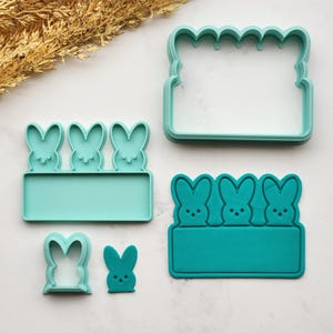 May include: A selection of teal cookie cutters in different sizes and shapes. The cutters feature bunny designs, ideal for Easter baking. Includes a large rectangular cutter with a scalloped top edge.