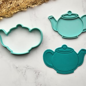 Può includere: Tre formine per biscotti a forma di teiera color teal. Uno è un contorno pieno e gli altri due sono pieni. Le formine sono su una superficie di marmo bianco e grigio, con una pianta secca nell'angolo in alto a sinistra.