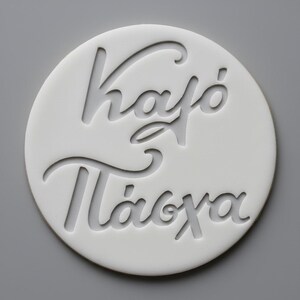 May include: A white, circular decorative piece with the Greek words "Καφό Πάσχα" cut out. The text is in a stylized, cursive font. The background is a solid gray.