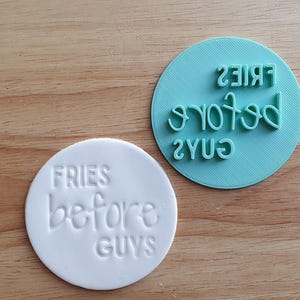 May include: A white circular stamp with the phrase "FRIES before GUYS" and a teal circular stamp with the phrase "FRIES before GUYS" in reverse. The stamps are on a wooden surface.