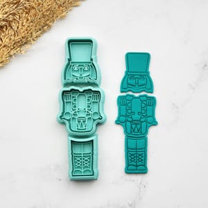 May include: Two teal plastic cookie cutters in the shape of a toy soldier. One is assembled, and the other is in three separate pieces. The soldier has a hat, a face, a drum, and boots. The cutters are on a white surface.
