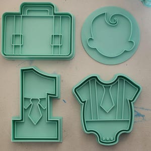 Könnte beinhalten: Eine Sammlung von mintgrünen Ausstechformen in verschiedenen Formen. Die Formen umfassen eine Aktentasche, ein Babygesicht, die Zahl eins mit Krawatte und einen Baby-Strampler mit Krawatte. Diese Ausstecher sind wahrscheinlich für thematische Kekse.