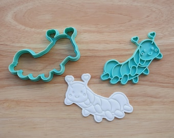 May include: Three caterpillar-shaped cookie cutters in the image. Two are teal, and one is white. The cutters have a cute design with heart-shaped antennae. The cutters are made of plastic and are on a wooden surface.