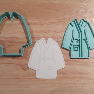 May include: Three cookie cutters shaped like a lab coat. One is a teal outline, another is a white solid shape, and the third is a teal lab coat with details. The cutters are on a wooden surface.
