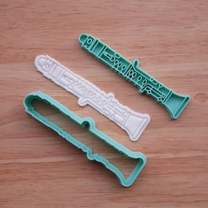 May include: Three clarinet-shaped cookie cutters in the image. One is white, one is teal, and the third is a teal outline. The cutters are made of plastic and are on a wooden surface. The design includes detailed instrument features.