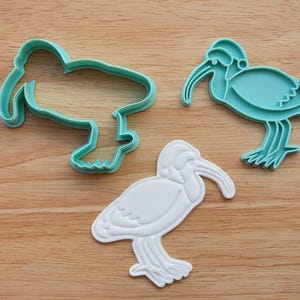 Puede incluir: Tres cortadores de galletas con forma de ibis. Uno es un contorno turquesa, otro es un ibis turquesa y el tercero es un ibis blanco. Los cortadores están sobre una superficie de madera. Ideales para hornear galletas temáticas.