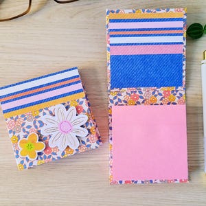 Puede incluir: Dos cuadernos con estampado floral, uno abierto y otro cerrado, con un bolígrafo blanco y dorado. El cuaderno abierto revela notas adhesivas rosas. Los cuadernos presentan diseños de margaritas y flores con detalles en azul, rosa y amarillo.