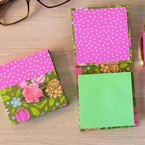 Puede incluir: Un bloc de notas con estampado floral y un bloc de notas cerrado a juego con una parte superior de lunares rosas. También se ven un bolígrafo rosa y gafas. El bloc de notas abierto tiene un bloc de notas adhesivas verdes en el interior. El estampado floral incluye rosas rosas y hojas verdes.