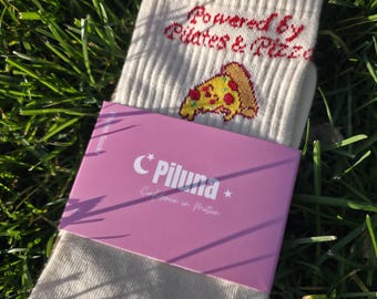 Puede incluir: Un par de calcetines de canalé color blanco roto con el texto bordado en rojo "Powered by Pilates & Pizza" sobre un pequeño gráfico de una porción de pizza. Los calcetines llevan una banda de papel rosa con el texto "Piluna" y "Confidence in Motion."