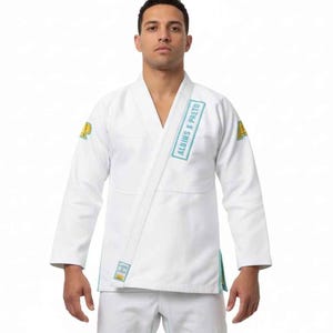 May include: White martial arts uniform with light blue accents. The uniform features the text "ALBINS S PRETO" on the shoulder and patches on the sleeves. The uniform is worn by a person.