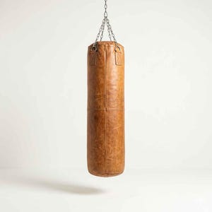 Könnte beinhalten: Ein brauner Leder-Boxsack, der an einer silbernen Kette hängt. Der zylindrische Sack weist Nähte und eine abgenutzte Textur auf. Die Oberseite hat zwei verstärkte Schlaufen für die Kette.
