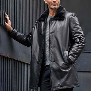 Könnte beinhalten: Ein schwarzer Ledermantel mit einem Kunstfellkragen. Der Mantel hat eine Knopfleiste, zwei Taschen und eine lange Länge. Die Person auf dem Bild trägt ein graues Hemd darunter.
