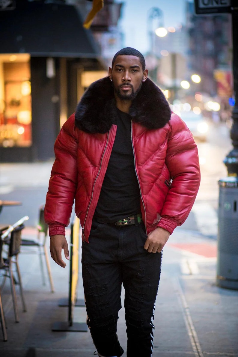 May include: A red quilted puffer jacket with a black fur collar. The person is wearing a black top, black jeans, and a camouflage belt. The jacket has a zip closure. The background shows a city street with buildings.
