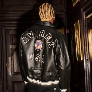 Könnte beinhalten: Schwarze Lederjacke mit weißem Besatz und dem Text "AVIREX USA" auf dem Rücken. Die Jacke hat einen Aufnäher mit der amerikanischen Flagge und den Worten "ALL AMERICANS 1975".