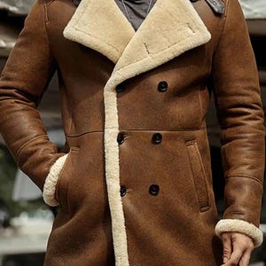 Könnte beinhalten: Ein brauner Ledermantel mit cremefarbenem Shearling-Kragen und -Futter. Der zweireihige Mantel hat schwarze Knöpfe, ein fallendes Revers und Taschen. Die Ärmel haben Shearling-Bündchen. Ein klassischer Wintermantel.