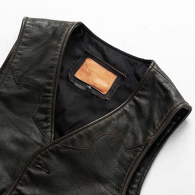 May include: Close-up of a black leather vest with a V-neck design. The vest has a button closure and decorative stitching along the edges. A brown leather label is sewn inside the vest. The vest is XL size.