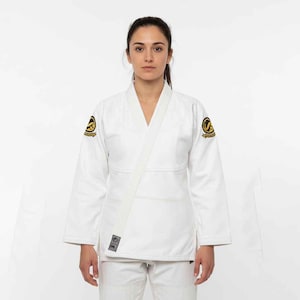 May include: White martial arts uniform with a V-neck top and matching trousers. The uniform has a white belt and patches with a gold and black design on the shoulders. The person is standing in front of a white background.