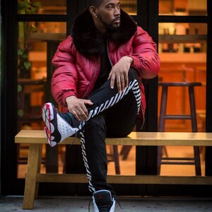 May include: A red puffer jacket with a black fur collar. The person is wearing black trousers with white stripes and black and white trainers with red accents. The person is sitting on a wooden bench.