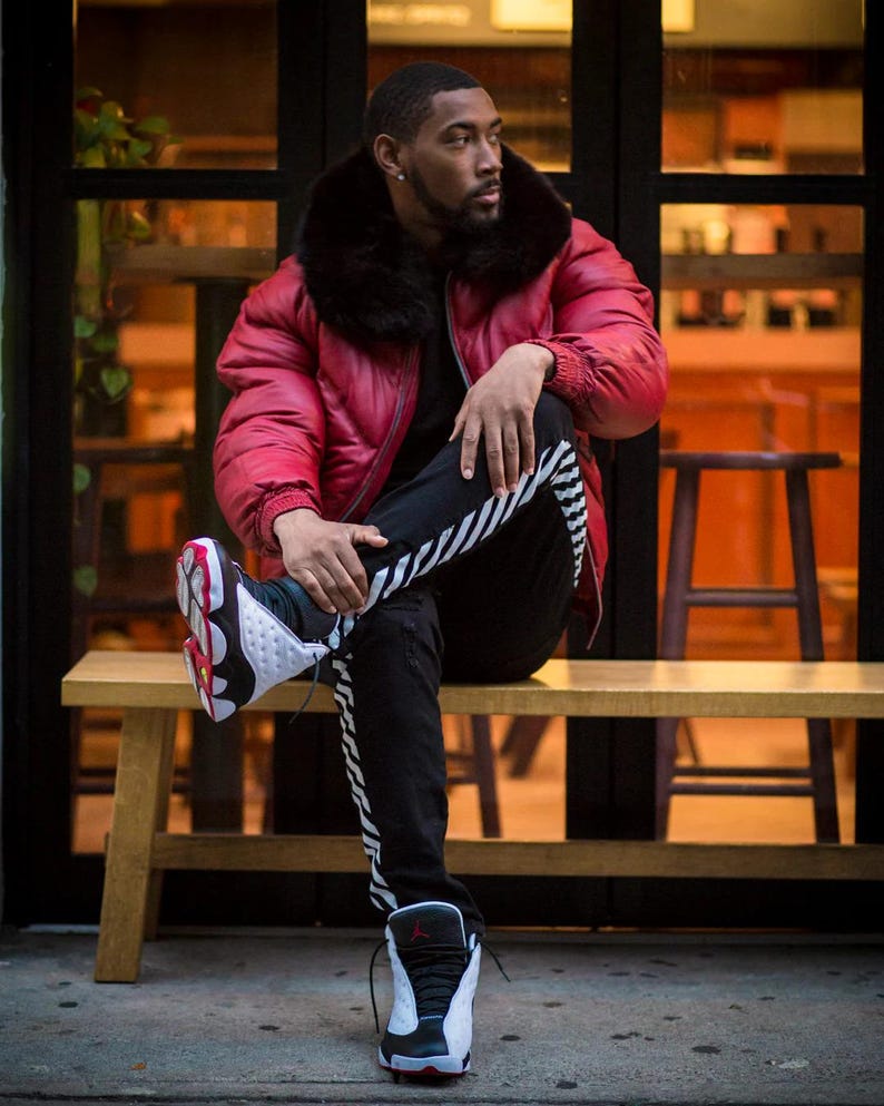 May include: A red puffer jacket with a black fur collar. The person is wearing black trousers with white stripes and black and white trainers with red accents. The person is sitting on a wooden bench.