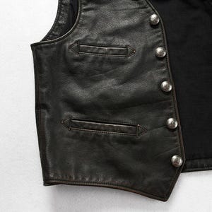 May include: Close-up of a black leather vest with two front pockets and silver-colored buttons. The vest has a classic design and appears to be well-worn, with visible texture and detailing. The vest is likely part of a biker or vintage-inspired collection.