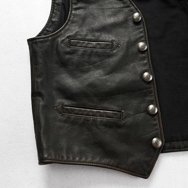 May include: Close-up of a black leather vest with two front pockets and silver-colored buttons. The vest has a classic design and appears to be well-worn, with visible texture and detailing. The vest is likely part of a biker or vintage-inspired collection.