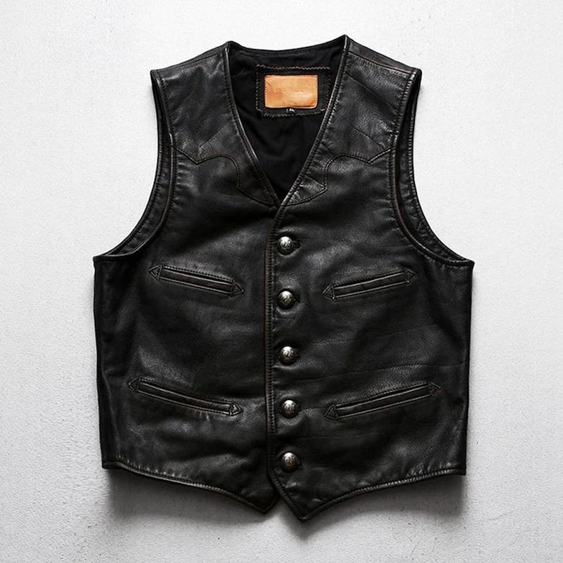 May include: A black leather vest with a V-neck and a button-up front. The vest has two front pockets and decorative stitching along the shoulders and chest. The vest has a small, rectangular patch on the back.