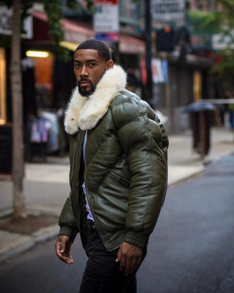 May include: Olive green puffer jacket with a cream-colored faux fur collar. The jacket has a quilted design and ribbed cuffs. The person is wearing a white shirt and black pants. The background is a city street.