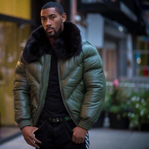 May include: An olive green puffer jacket with a black fur collar. The jacket has a quilted design and a zipper closure. The person is wearing a black shirt and black pants with a camouflage belt.