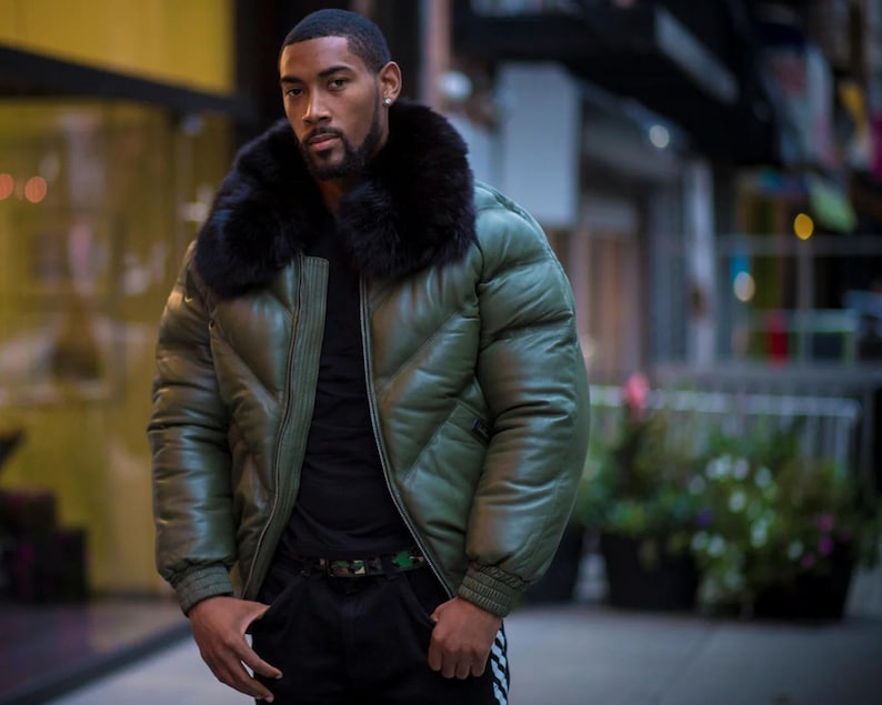 May include: An olive green puffer jacket with a black fur collar. The jacket has a quilted design and a zipper closure. The person is wearing a black shirt and black pants with a camouflage belt.