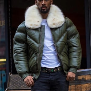 May include: An olive green bomber jacket with a white fur collar. The jacket has a quilted design and a zipper closure. The person is wearing a white t-shirt and black jeans. A camouflage belt is visible.