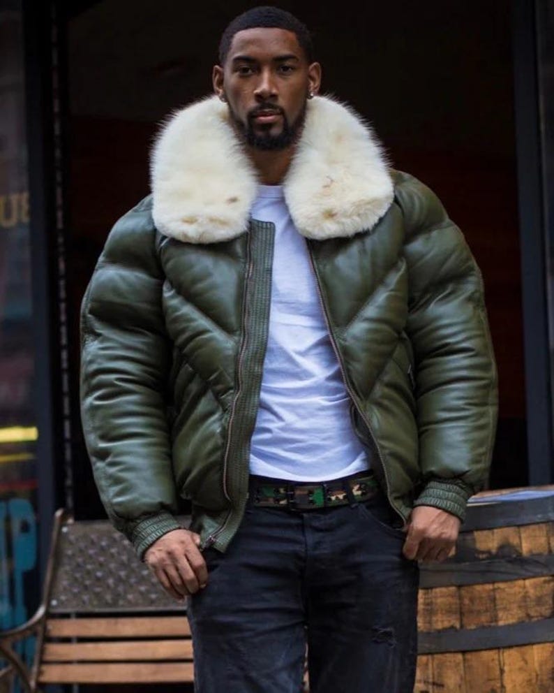 May include: An olive green bomber jacket with a white fur collar. The jacket has a quilted design and a zipper closure. The person is wearing a white t-shirt and black jeans. A camouflage belt is visible.