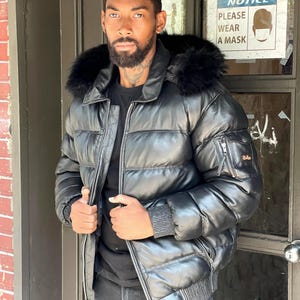 Könnte beinhalten: Schwarze Leder-Steppjacke mit schwarzer, fellgefütterter Kapuze. Die Jacke hat einen Reißverschluss, mehrere Taschen und gerippte Bündchen. Ein Schild im Hintergrund lautet "NOTICE PLEASE WEAR A MASK".