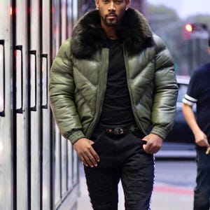 May include: An olive green puffer jacket with a black fur collar. The jacket has a quilted design and a zipper closure. The person is wearing a black shirt and black ripped jeans with white stripes. A camouflage belt is visible.