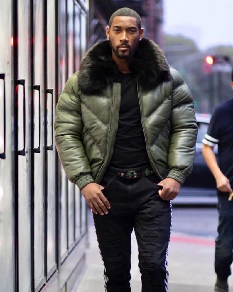 May include: An olive green puffer jacket with a black fur collar. The jacket has a quilted design and a zipper closure. The person is wearing a black shirt and black ripped jeans with white stripes. A camouflage belt is visible.