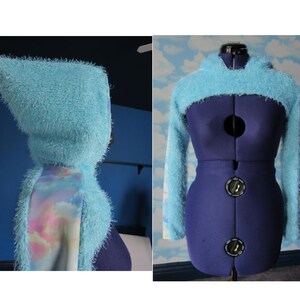May include: A light blue fuzzy hooded shrug with a rainbow cloud print on the back. The shrug is shown on a mannequin.