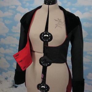 May include: A black cropped jacket with a red lining and velvet collar. The jacket has a button closure and is displayed on a mannequin.