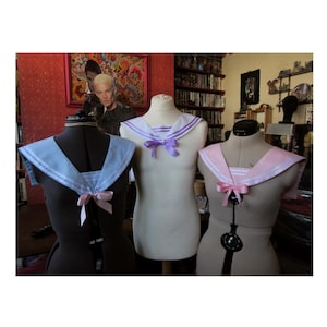 May include: Three sailor collar tops in pastel colors displayed on dress forms. The collars are light blue, white with purple stripes, and pink, each with a matching bow. The background includes a painting and a bookshelf.